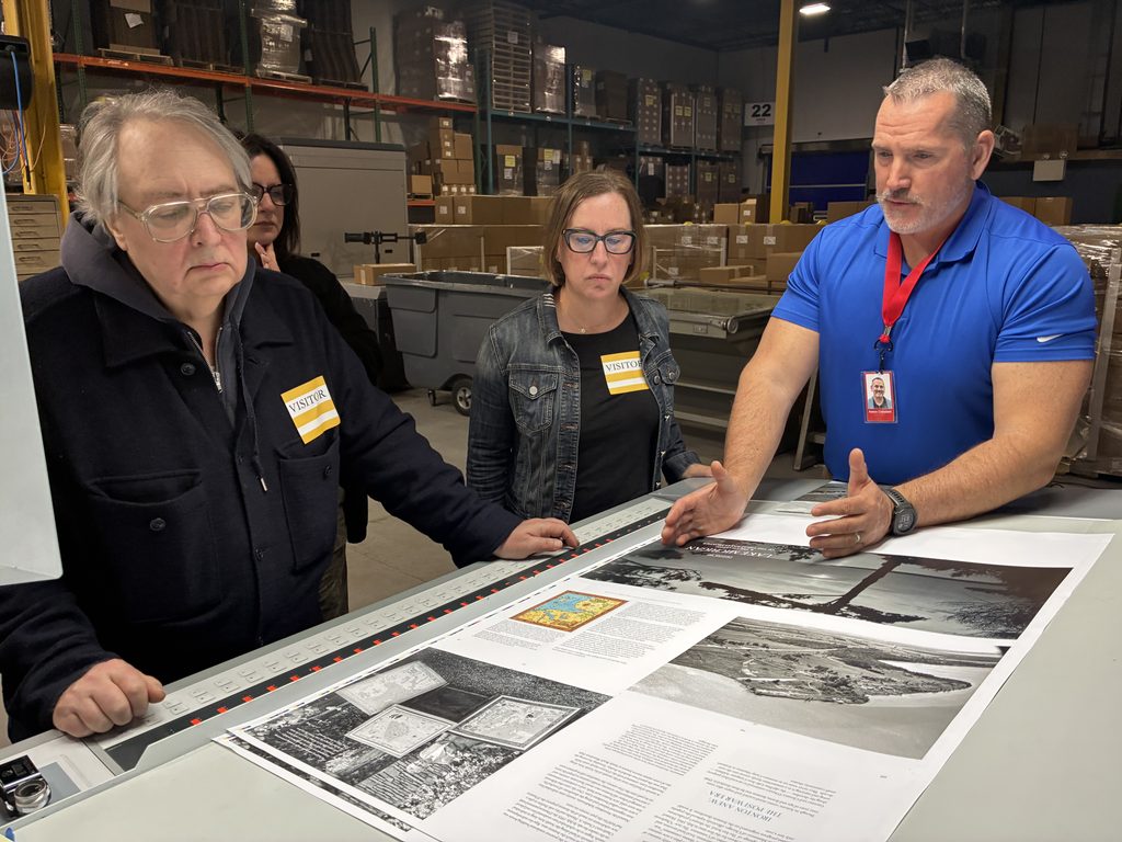 A man at Puritan Capital, a printing house, shows a team of visitors working on "A City on Three Lakes" a large, printed layout. The sheet appears about 3 feet tall by two feet wide and contains a mix of photos and blocks of text.