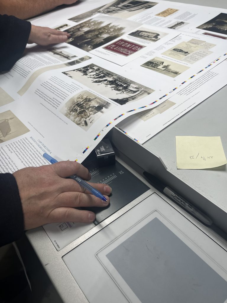 A long page of printed photographs being reviewed. A hand with a pen, as well as a sticky note with written measurements, are also visible.