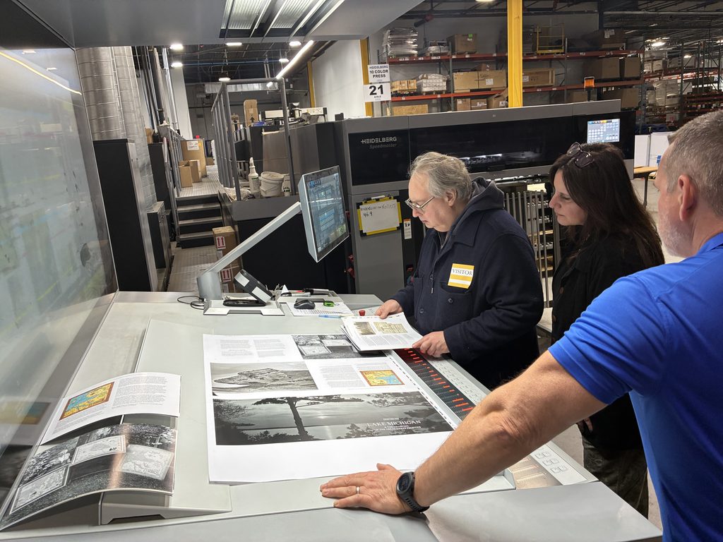 Member of the team creating "A City on Three Lakes" and an employee from the printing house Puritan Capital review a large, printed layout. Machines, tools, and various equipment can be seen in the background.